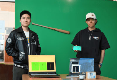 two young men on behind a table with two laptops, one wearing a white baseball cap, both in front of a green screen with a baseball bat hanging in front of it.